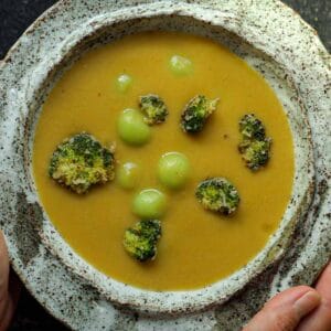 A rustic ceramic bowl brimming with creamy broccoli soup, adorned with roasted florets and green dumplings. Hands gently cradle the bowl, emphasizing the earthy texture of the pottery.